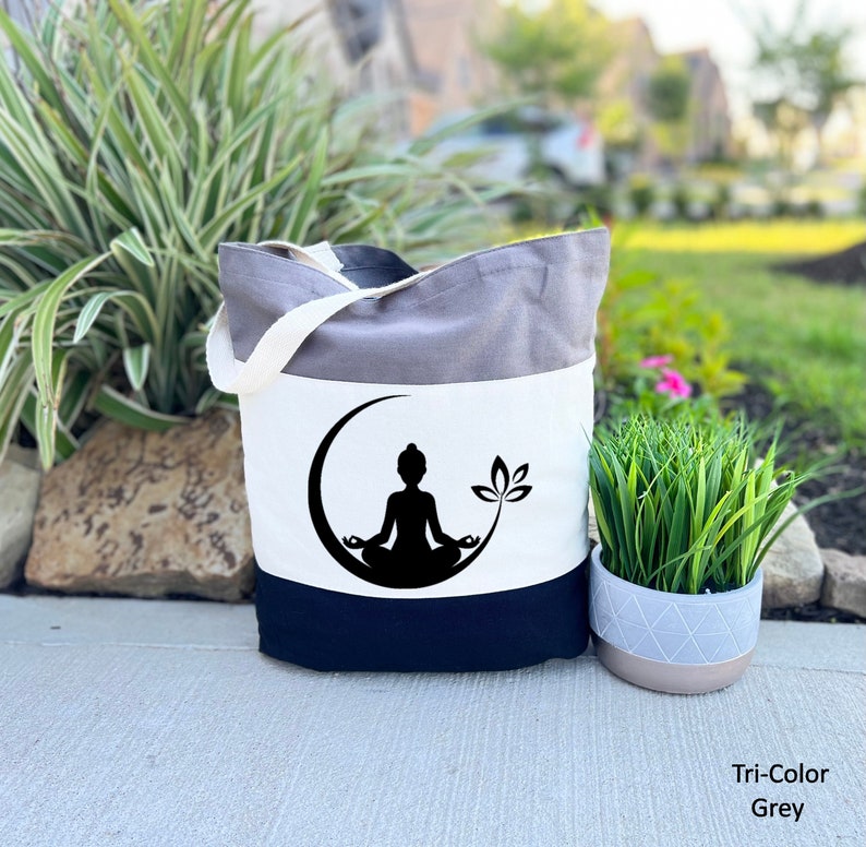 May include: A tri-color tote bag with a grey, white, and black design. The bag features a black silhouette of a person meditating in a crescent moon with a lotus flower.