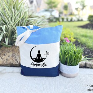 May include: A tri-color tote bag with a blue, white, and navy blue design. The bag features a black silhouette of a person meditating in a crescent moon with a lotus flower. The name "Amanda" is printed in black below the image.