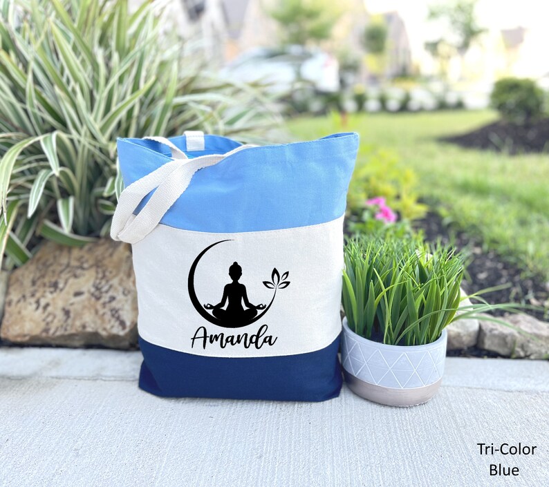 May include: A tri-color tote bag with a blue, white, and navy blue design. The bag features a black silhouette of a person meditating in a crescent moon with a lotus flower. The name "Amanda" is printed in black below the image.