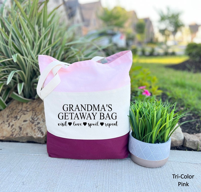 Pu&ograve; includere: Una borsa tote tricolore con un design rosa, bianco e bordeaux. La borsa ha il testo "Grandma's Getaway Bag" e "visit love spoil repeat" stampato su di essa.