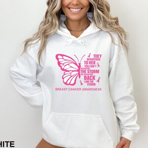 May include: White hooded sweatshirt with a pink butterfly graphic and the text "They whispered to her you can't withstand the storm she whispered back I am the storm Breast Cancer Awareness".