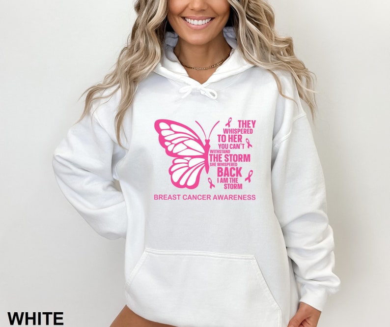 May include: White hooded sweatshirt with a pink butterfly graphic and the text "They whispered to her you can't withstand the storm she whispered back I am the storm Breast Cancer Awareness".