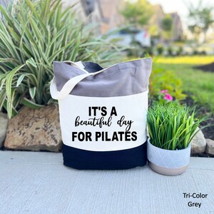 May include: A tri-color grey tote bag with a white strap. The bag has a grey top, a white middle section, and a navy blue bottom. The text "IT'S A beautiful day FOR PILATES" is printed in black on the white section.
