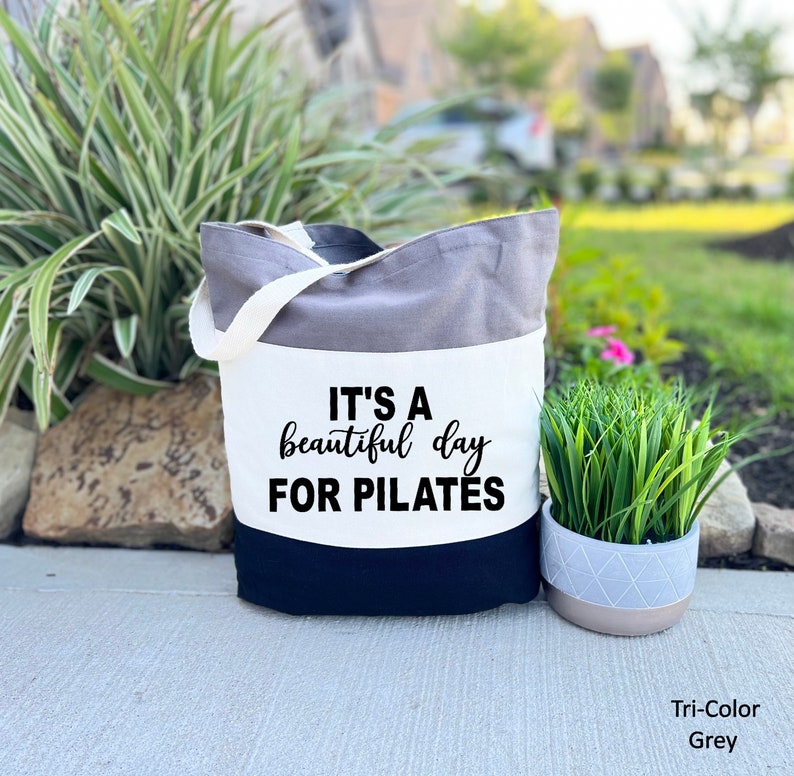 May include: A tri-color grey tote bag with a white strap. The bag has a grey top, a white middle section, and a navy blue bottom. The text "IT'S A beautiful day FOR PILATES" is printed in black on the white section.