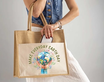 May include: A beige jute tote bag with a front pocket featuring the text "MAKE EVERYDAY EARTH DAY" around a graphic of hands holding the Earth surrounded by flowers. The bag has tan handles and is being held by a person.