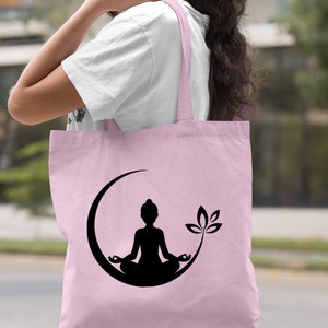 May include: A light pink tote bag with a black silhouette of a person meditating in a crescent moon with a lotus flower. The bag has black handles.