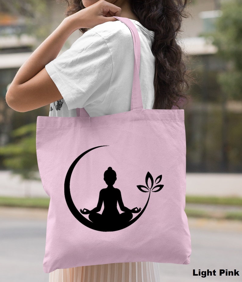 May include: A light pink tote bag with a black silhouette of a person meditating in a crescent moon with a lotus flower. The bag has black handles.