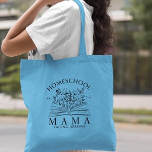 Peut inclure: Sac fourre-tout bleu clair avec l'inscription "Homeschool Mama Raising Arrows" en noir. Le motif comprend un livre ouvert avec des illustrations florales et de papillons. Le sac est port&eacute; sur l'&eacute;paule.
