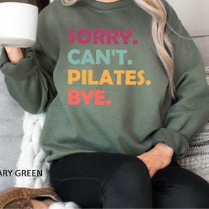 May include: A green sweatshirt with the text "SORRY. CAN'T. PILATES. BYE." in colorful letters. The sweatshirt is being worn by a person sitting on a couch.