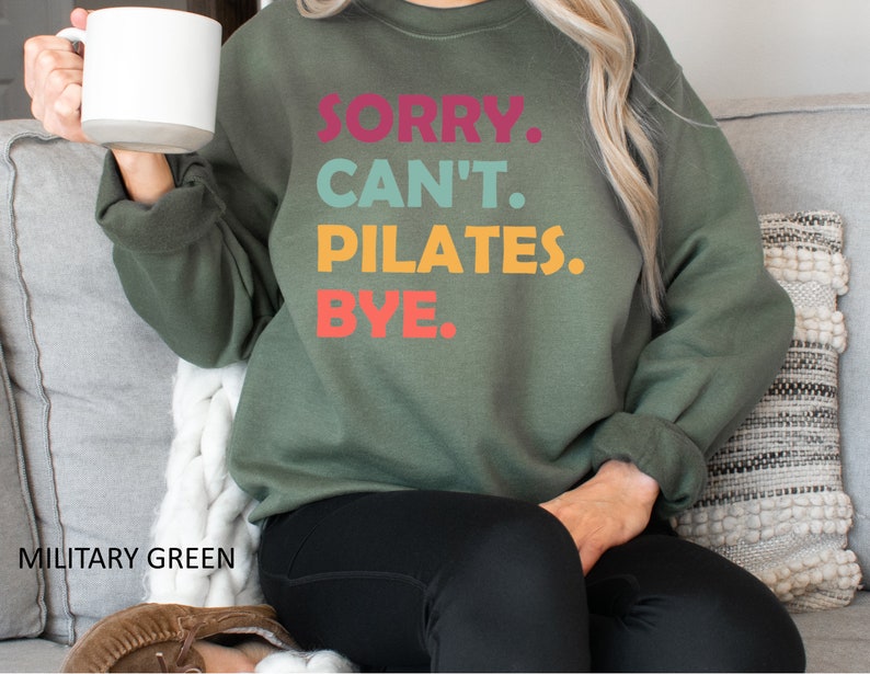 May include: A green sweatshirt with the text "SORRY. CAN'T. PILATES. BYE." in colorful letters. The sweatshirt is being worn by a person sitting on a couch.
