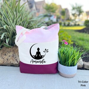 May include: A tri-color tote bag with a pink, white, and burgundy design. The bag features a black silhouette of a person meditating on a crescent moon with a lotus flower. The name "Amanda" is printed in black below the image.