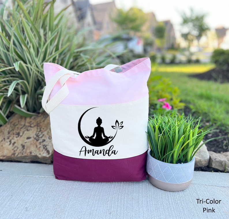 May include: A tri-color tote bag with a pink, white, and burgundy design. The bag features a black silhouette of a person meditating on a crescent moon with a lotus flower. The name "Amanda" is printed in black below the image.