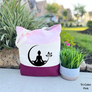 May include: A tri-color tote bag with a pink, white, and burgundy design. The bag features a black silhouette of a person meditating in a crescent moon with a lotus flower.