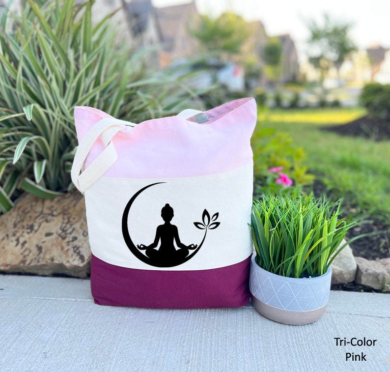 May include: A tri-color tote bag with a pink, white, and burgundy design. The bag features a black silhouette of a person meditating in a crescent moon with a lotus flower.