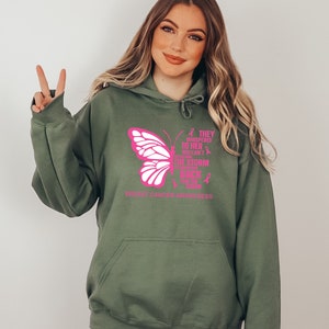 May include: A green hooded sweatshirt with a pink butterfly graphic and the text "They whispered to her you can't withstand the storm but she came back stronger than the storm" and "Breast Cancer Awareness".