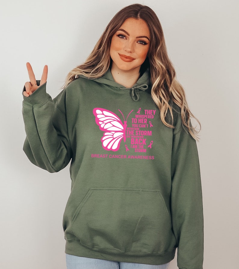 May include: A green hooded sweatshirt with a pink butterfly graphic and the text "They whispered to her you can't withstand the storm but she came back stronger than the storm" and "Breast Cancer Awareness".