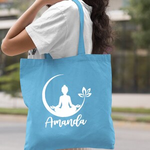 May include: Light blue tote bag with a white silhouette of a person meditating on a crescent moon with a lotus flower. The bag has the name "Amanda" printed below the image.