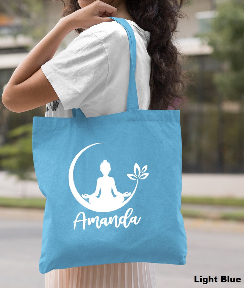 May include: Light blue tote bag with a white silhouette of a person meditating on a crescent moon with a lotus flower. The bag has the name "Amanda" printed below the image.