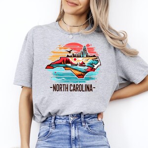 May include: A gray t-shirt with a graphic of the state of North Carolina in a sunset color palette. The graphic includes a cityscape, mountains, and a lake. The text "North Carolina" is printed below the graphic.