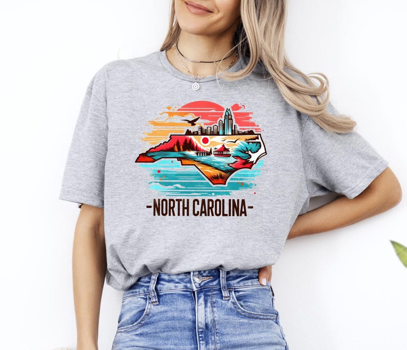 May include: A gray t-shirt with a graphic of the state of North Carolina in a sunset color palette. The graphic includes a cityscape, mountains, and a lake. The text "North Carolina" is printed below the graphic.