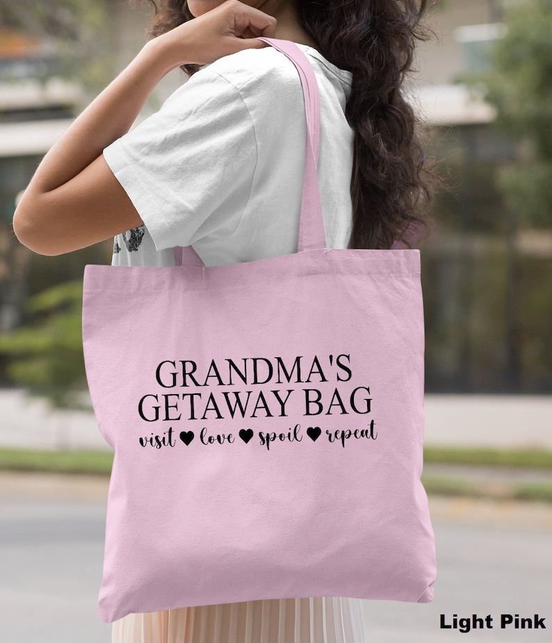 Pu&ograve; includere: Una borsa tote rosa chiaro con il testo "Grandma's Getaway Bag visit love spoil repeat" stampato in nero. La borsa ha manici neri.