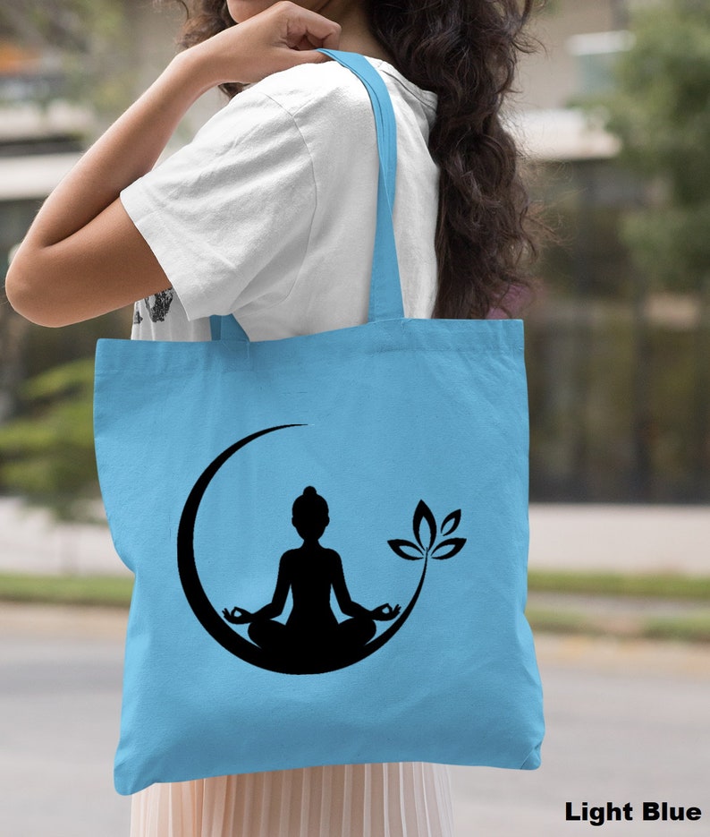 May include: Light blue tote bag with a black silhouette of a person meditating in a crescent moon with a lotus flower.