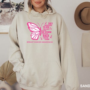 May include: A light beige hoodie with a pink butterfly graphic and the text "They whispered to her you can't withstand the storm she whispered back I am the storm" and "Breast Cancer Awareness".