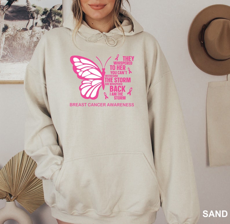 May include: A light beige hoodie with a pink butterfly graphic and the text "They whispered to her you can't withstand the storm she whispered back I am the storm" and "Breast Cancer Awareness".