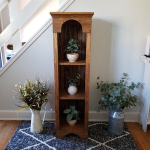 May include: A wooden shelf unit with three shelves, each holding a potted plant. The shelf unit is stained a dark brown color and has a decorative arched top. The plants are all green and leafy.