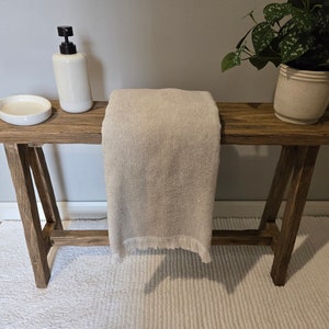 May include: A rustic wooden table with a white towel draped over it. The table has two legs on each side and is sitting on a white rug. There is a white soap dispenser and a white dish on the table.