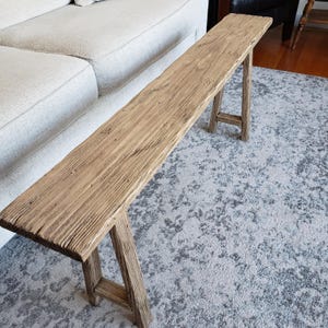 May include: A rustic wooden bench with a natural brown finish and a distressed look. The bench has a long, rectangular top and two simple, angled legs. The bench is sitting on a gray and white patterned rug.