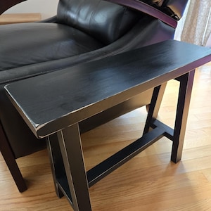 May include: A small, black wooden side table with a rectangular top and angled legs. The table has a distressed finish, showing hints of the wood grain. The table is designed to be placed next to a chair or sofa.