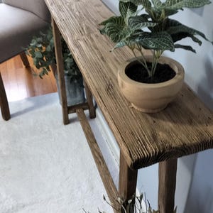 May include: A rustic, wooden console table with a natural finish. A small potted plant with green and white leaves sits on the table. The table has a narrow design and is placed against a light blue wall.