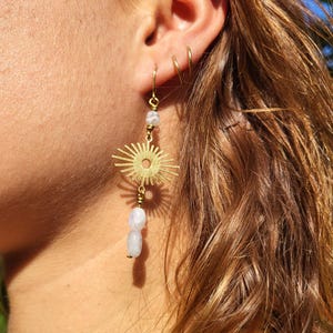 Könnte beinhalten: Goldfarbene Hängeohrringe. Jeder Ohrring hat ein Sonnenstrahl-Design, kleine weiße Perlen und eine größere weiße Perle unten. Die Ohrringe hängen am Ohr einer Person.