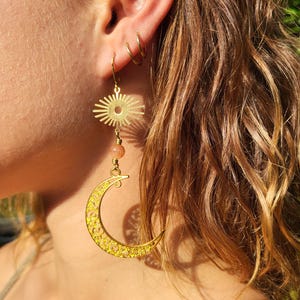 Könnte beinhalten: Goldfarbene Ohrringe mit einem Sonnenmuster, einer pfirsichfarbenen Perle und einem Mond mit aufwendigen Ausschnitten. Die Ohrringe hängen an einem goldfarbenen Ohrhaken. Die Ohrringe sind Schmuck mit Himmelsmotiven.