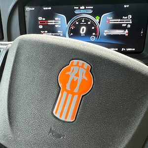 May include: A black leather steering wheel with a silver and orange Peterbilt logo. The logo features a stylized image of a woman walking. The steering wheel is in a truck cab with a digital dashboard displaying various readings, including speed, fuel level, and temperature.