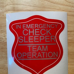 May include: A red and white sticker featuring the text "IN EMERGENCY CHECK SLEEPER TEAM OPERATION" in silver lettering. The sticker is shield-shaped.