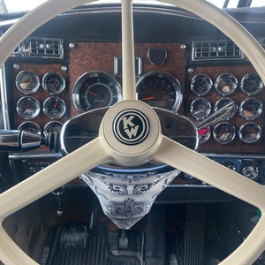 May include: A close-up view of a beige steering wheel with a black and white bandana hanging from the bottom. The steering wheel has a chrome center with a black and white KW logo. The dashboard behind the steering wheel has a woodgrain finish and several gauges.