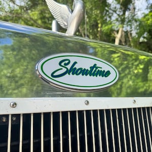 May include: Close-up of a vintage vehicle's front. A chrome bird hood ornament sits above an oval emblem with the word "Showtime" in green script. The vehicle's grill is white with vertical black bars.