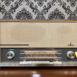 Könnte beinhalten: Vintage Siemens Radio mit hellbeigem Lautsprecherbezug und dunkelgrauem Bedienfeld. Das Radio hat einen Holzrahmen und silberne Knöpfe. Das Siemens-Logo ist sichtbar. Das Radio steht auf einer dunkelbraunen Oberfläche.