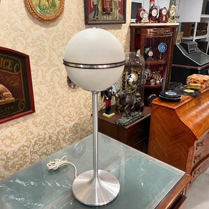 May include: A white globe lamp with a silver base and a silver band around the middle of the globe. The lamp is on a glass table top.