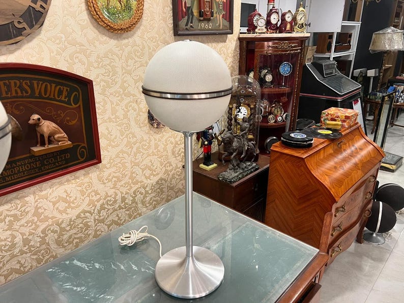 May include: A white globe lamp with a silver base and a silver band around the middle of the globe. The lamp is on a glass table top.