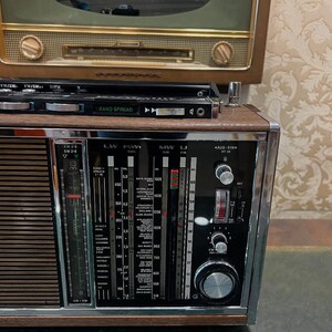 May include: Vintage wooden radio with a silver and black face, featuring dials, a tuning scale, and a speaker grill. The radio has a retro design, with a second, older radio visible in the background. The radio has the words "BAND SPREAD" on the top.