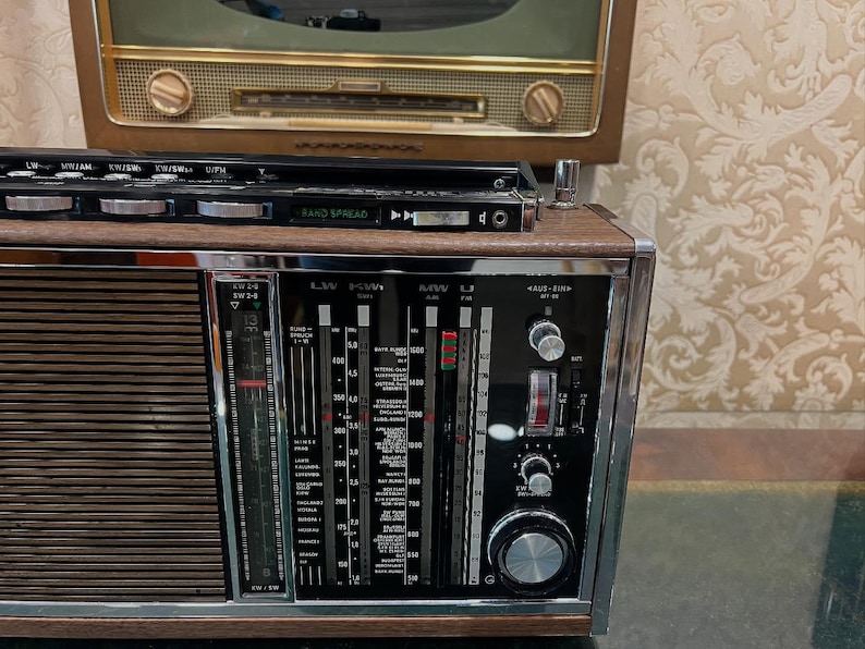 May include: Vintage wooden radio with a silver and black face, featuring dials, a tuning scale, and a speaker grill. The radio has a retro design, with a second, older radio visible in the background. The radio has the words "BAND SPREAD" on the top.
