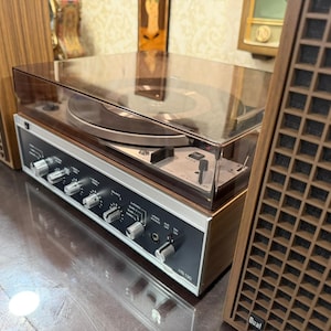 May include: A white ceramic mug with a black handle and the text "But First, Coffee" in a bold, black font. The mug is set against a blurred background, suggesting a cozy setting.