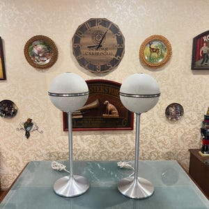 May include: Two vintage white and silver mid-century modern speakers on silver stands. The speakers have a round shape with a silver grill and a white fabric covering. The stands are tall and slender with a round base.
