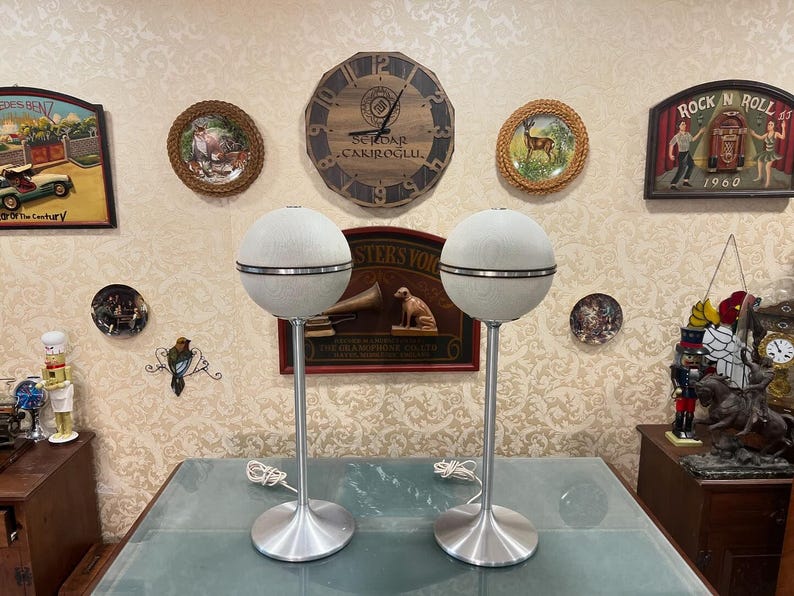 May include: Two vintage white and silver mid-century modern speakers on silver stands. The speakers have a round shape with a silver grill and a white fabric covering. The stands are tall and slender with a round base.