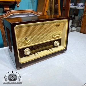 Op de afbeelding: Een vintage houten radio met een gouden luidsprekerrooster en witte knoppen. De radio heeft een bruine houten behuizing en een witte voorplaat met een zwarte wijzerplaat en een witte afstemknop.