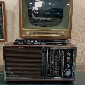 May include: A vintage Grundig radio and television set. The radio is brown with a silver trim and a speaker grill. The television has a wooden frame and a screen displaying an image of an airplane. A small black component sits between the two.