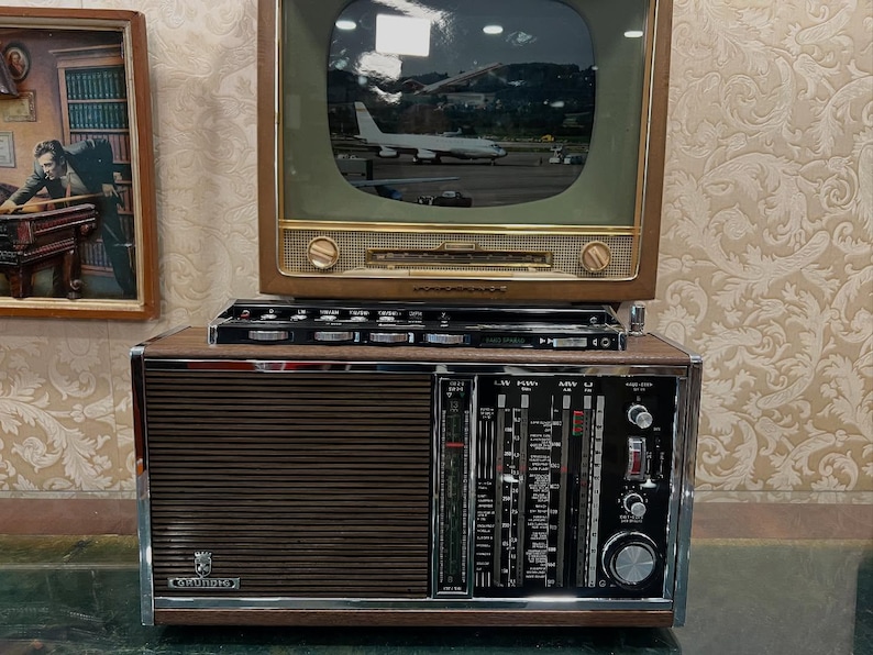 May include: A vintage Grundig radio and television set. The radio is brown with a silver trim and a speaker grill. The television has a wooden frame and a screen displaying an image of an airplane. A small black component sits between the two.